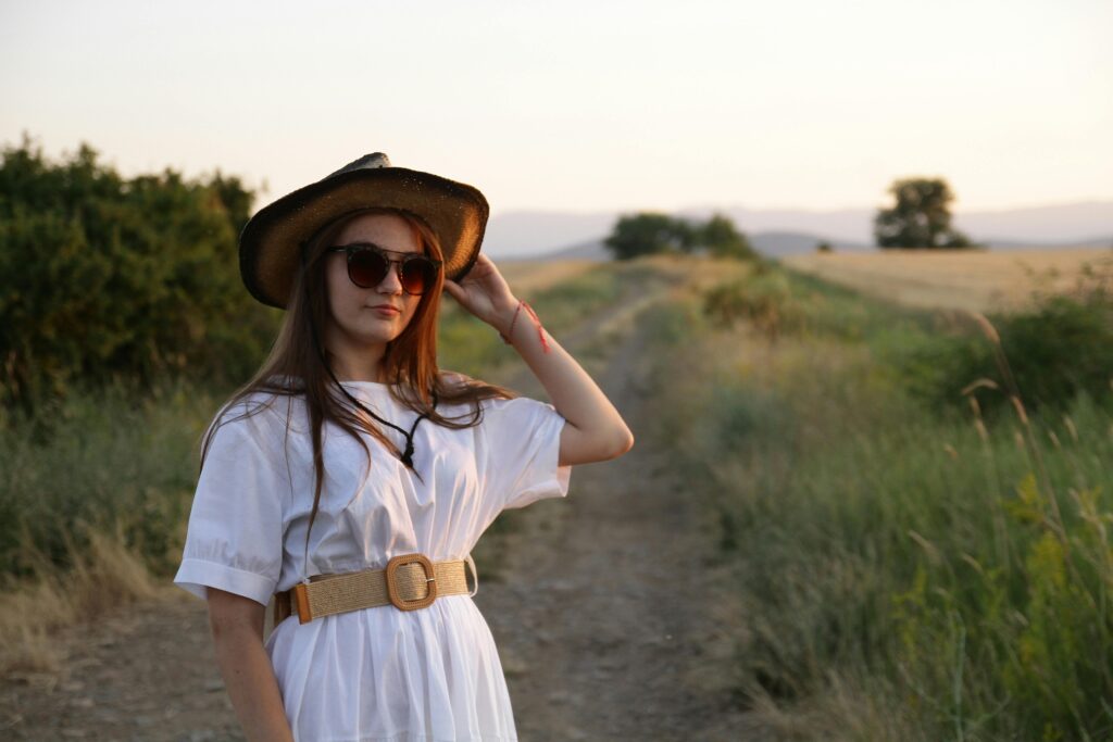 A young woman in sunglasses and a straw hat poses on a rural path at sunset.